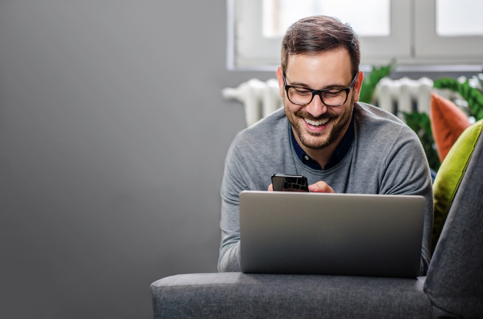 Man sat on a sofa with his laptop smiling at his phone